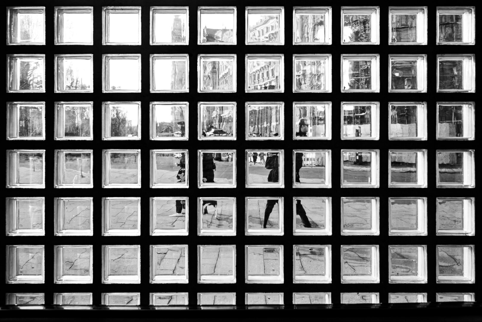 The image is a black and white, overhead shot of a wall constructed from glass blocks. The blocks are arranged in a grid of five rows and eight columns, with a few extra blocks at the bottom row appearing to be slightly different in size and orientation. The overall effect is abstract and geometric, with the varying light and reflections within each block creating a textured, fragmented view of the outside world.