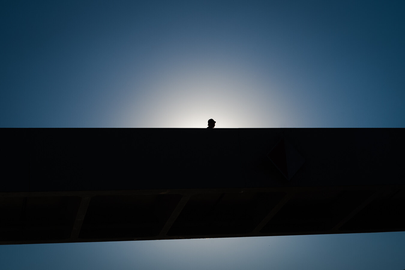 The image features a minimalist composition dominated by dark silhouettes against a bright, gradient sky. The main subject is the silhouette of a person's head and shoulders, seen from the side, peeking over the top edge of a large, dark structure. The person appears to be wearing a hat or hood. This structure, also in silhouette, runs horizontally across the lower half of the frame, dividing the image into distinct sections.