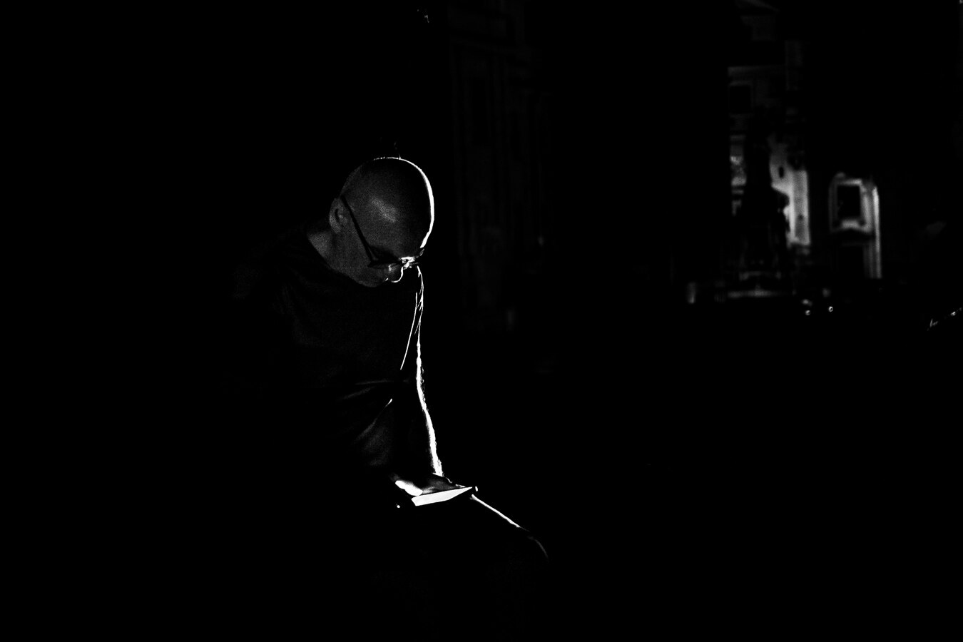 The image is a high-contrast, black and white, medium shot of a bald man with glasses, who is looking down at the glowing screen of his phone held in his hands. The man is positioned on the left side of the frame, with the majority of the image being consumed by darkness.
