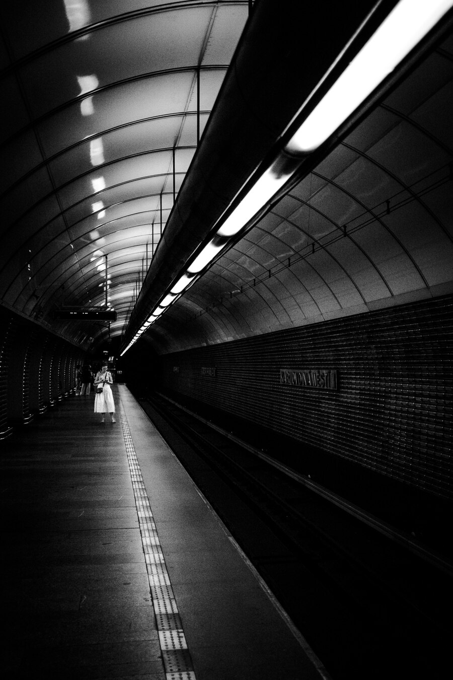 The image depicts a dramatic, high-contrast black and white scene within a subway station. The perspective is from a low angle, looking down a long, arched tunnel.

The primary elements are the architectural features of the station and a solitary figure. The ceiling is a prominent feature, characterized by its curved, ribbed structure and illuminated by long, fluorescent tube lights.  
In the middle ground, on the left side of the frame, a person stands on the platform. She is dressed in a light-colored, flowing garment, which stands out against the dark surroundings.