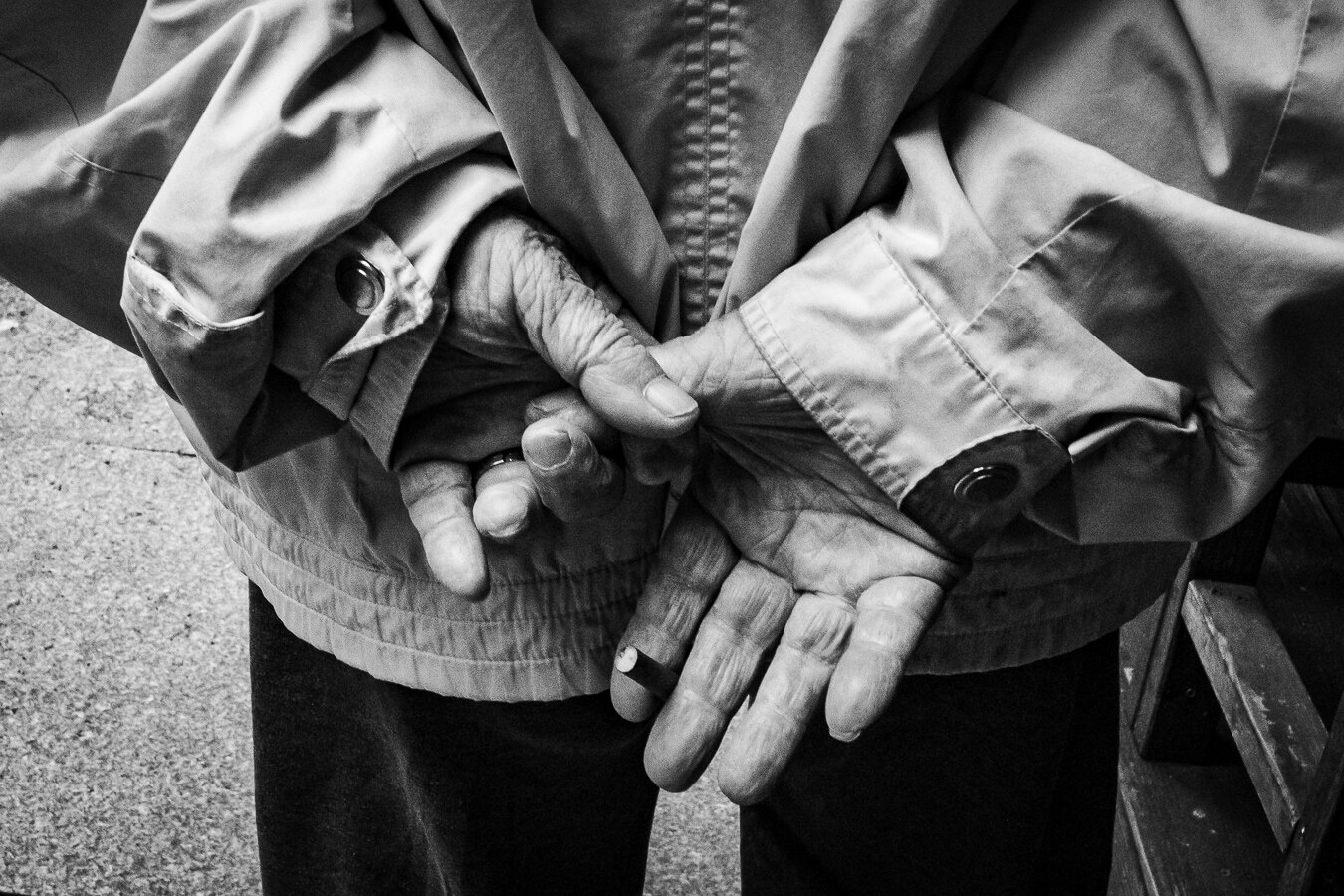 The image is a close-up, black and white photograph focusing on a person's hands clasped behind their back. The subject is wearing a light-colored jacket with visible stitching and buttons. The hands appear to belong to an elderly individual, as evidenced by the wrinkled skin and prominent veins. One hand wears a ring on the ring finger. The texture of the hands is detailed, highlighting the signs of age. The composition is tight, with the hands and jacket filling most of the frame. The background is blurred, with a speckled texture on the left, possibly concrete, and a dark, indistinct area on the right that might be furniture or clothing. The lighting creates strong shadows and highlights, emphasizing the contours of the hands and the folds of the jacket.