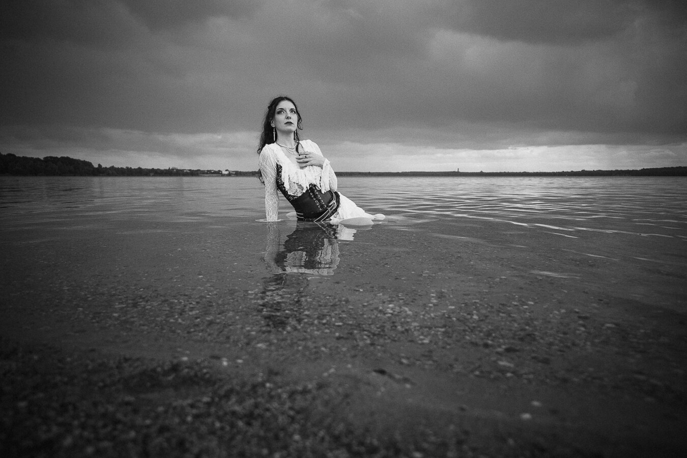 The image is a black and white, low-angle shot of a woman partially submerged in shallow water. She is positioned on the right side of the frame, leaning back and looking upwards and to her left. Her body is angled, with her right leg bent and submerged, and her left leg extended slightly. She is wearing a white, flowing dress with lace detailing around the neckline and cuffs, and a dark, structured corset over it. The corset has visible lacing and decorative elements. Her hand is placed over her chest, on top of the lace trim of her dress. She has dark hair pulled back, and noticeable earrings.