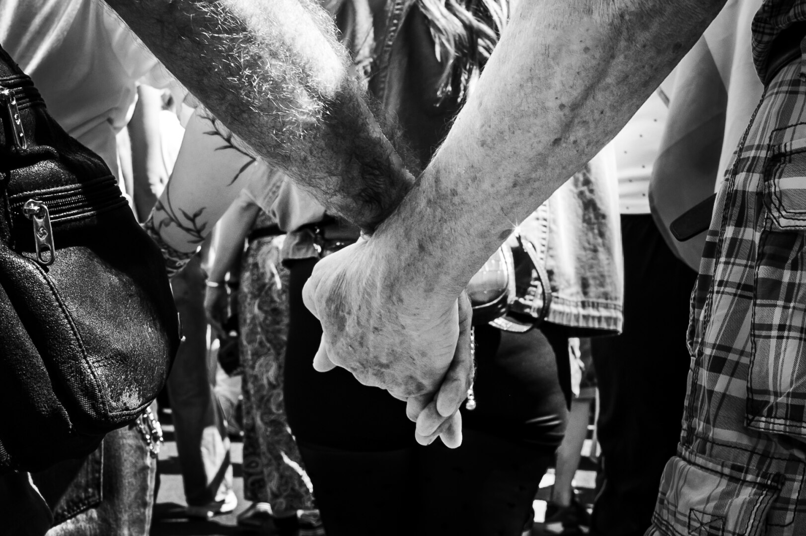 The image is a black and white, close-up photograph focusing on the hands of two people who appear to be in a crowd or gathering. The dominant element is the clasped hands, positioned in the center of the frame. One hand, belonging to a person with a more wrinkled and age-spotted skin tone, is clasped firmly by another hand.