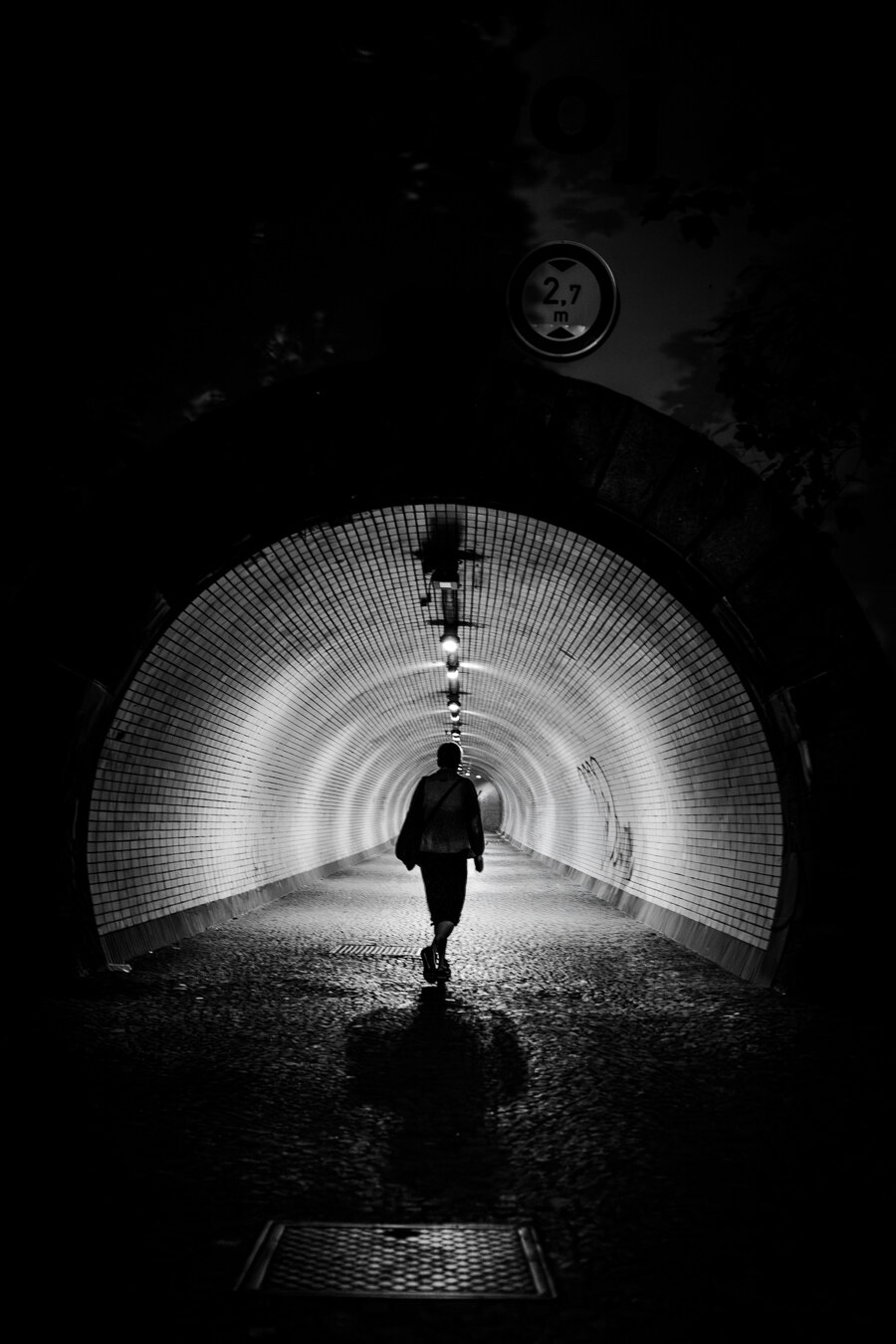 The image is a black and white photograph depicting a person walking away from the viewer into a tunnel. The person in the image is silhouetted against the light, making their features indistinguishable. The tunnel's interior is illuminated by a series of lights hanging from the ceiling, creating a strong sense of perspective and leading the eye deeper into the darkness. The walls of the tunnel are lined with small, square tiles, which reflect the light and create a textured, almost shimmering effect. The floor of the tunnel appears to be paved with cobblestones.