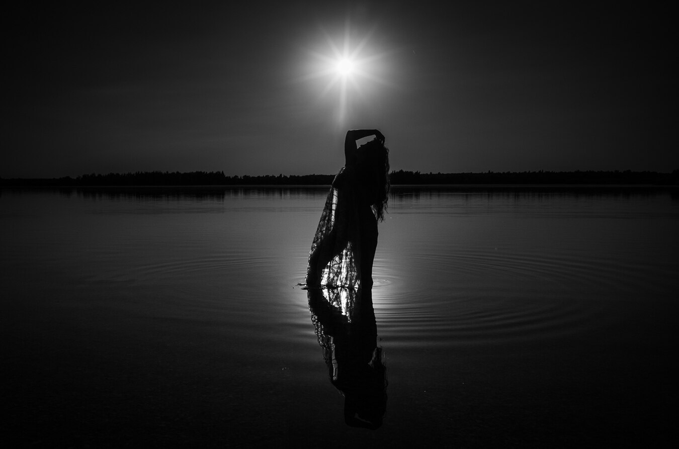 The image is a dramatic black and white photograph captured at sunset. The scene is dominated by a vast expanse of calm water, with a dark, silhouetted treeline forming the horizon in the distance. Above the treeline, the sky is a gradient of dark grey to black.

The main focal point is a solitary female figure standing in the water, silhouetted against the bright, starburst-like sun. The sun is positioned almost directly overhead and slightly forward of the figure, its intense light creating a powerful lens flare effect. The figure is facing away from the viewer, with one arm raised to the back of her head, and her body is slightly curved. She appears to be wearing a flowing, possibly sheer garment that drapes around her and extends into the water. The fabric has a textured pattern that is partially illuminated by the sun, creating intricate light and shadow play. Her hair is long and appears to flow loosely.