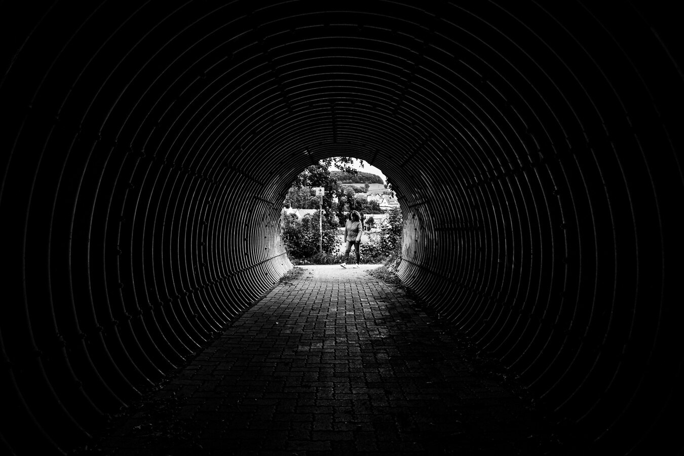 The image is a black and white, low-angle shot looking through a dark, corrugated metal tunnel towards an opening. The tunnel's circular structure dominates the foreground and midground, creating a strong sense of depth and leading the eye outwards. The texture of the corrugated metal is emphasized by the dramatic lighting, with highlights catching the ridges and shadows deepening the grooves. The floor of the tunnel is paved with dark bricks, which appear wet or reflective in places, adding to the moody atmosphere.

At the end of the tunnel, the opening reveals a bright, outdoor scene. In the center of this opening stands a person, silhouetted against the brighter background.